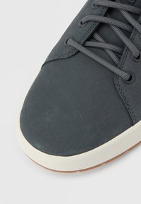 Pelēks sporta apavu ar gludu augšdaļu, auklu dizainu, kontrastējošu balto zoli un gaiši brūnu gumijas zolījumu.