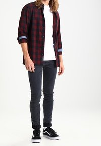 Camisa de cuadros roja y negra con botones sobre una camiseta blanca, combinada con jeans de mezclilla oscura y zapatillas negras con detalles blancos.