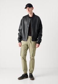Zwarte neplederen bomberjack, olijfgroene cargobroek en zwarte sneakers. Pet heeft een logo. Slim fit; jack heeft gewatteerde details en ritsluiting.