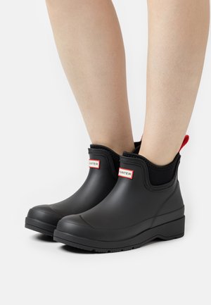 Bottines en caoutchouc noir, avec une texture lisse, un bout rond et une languette rouge. Présente une étiquette avec logo près du haut.