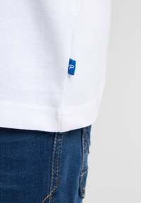 Bílá bavlněná tričko s modrou cedulkou na lemu, spárované s tmavě modrými denimovými džíny. Viditelné švy a čisté okraje.