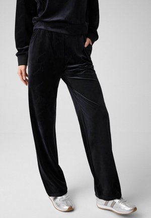 Persona con pantalones anchos de terciopelo negro y zapatillas plateadas, de pie con una mano en el bolsillo frente a un fondo liso.