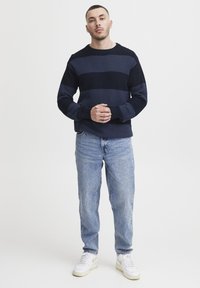 Pulover tricotat cu dungi navy și albastru închis, textură ribbed, guler rotund, asortat cu blugi denim de un albastru deschis și adidași albi.