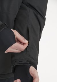 Svart vattentät jacka med en åtsittande mudd och justering med kardborreband. En hand justerar en sidoficka på ärmen, vilket visar på ett slätt och hållbart material.