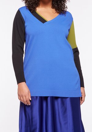 Vrouw met een blauwe V-hals trui met zwarte en olijfgroene colourblocking op de lange mouwen en een glanzende blauwe rok.