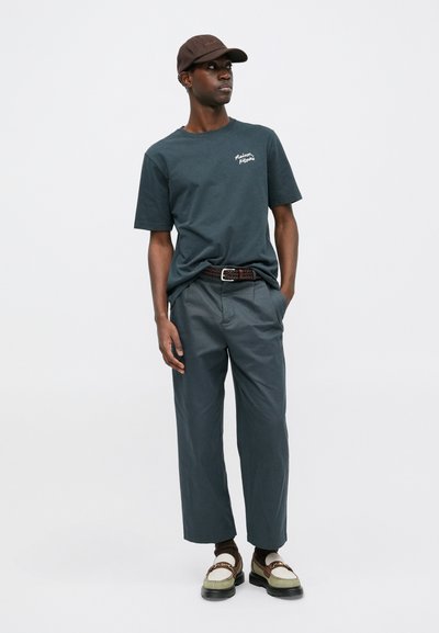 Dunkelblaues Baumwoll-T-Shirt mit kurzen Ärmeln, graue Hosen, brafter Gürtel und mehrfarbige Schuhe. Braune Mütze mit Logodetails.