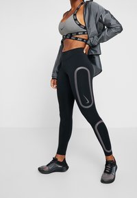 Žena v černých legínách Nike s šedými oválnými vzory, šedém sportovním podprsenku, šedé bundě a černých běžeckých botách, stojící na bílém pozadí.