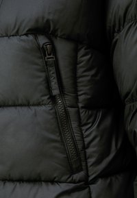 Jachetă puffer neagră, cu un buzunar cu fermoar vizibil, textură matlasată și un finisaj neted. Materialul este ușor și izolat.