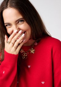 Pull rouge avec des poignets côtelés, petits motifs de cœur, chaîne en or avec des charms floraux et de trèfle, et des ongles soigneusement manucurés.