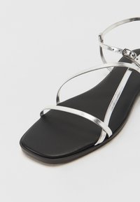 Sorte flate sandaler med metalliske sølv krossremmer, firkantet tåform, glatt matt tekstur og spenneaccent på ankelremmen.