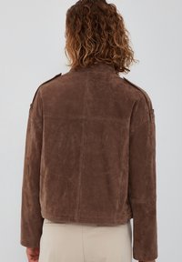 Bruine suède jas met een ontspannen pasvorm, voorzien van een hoge kraag, schouderepauletten en een gestructureerd oppervlak. Minimalistisch ontwerp, geen zichtbare hardware.