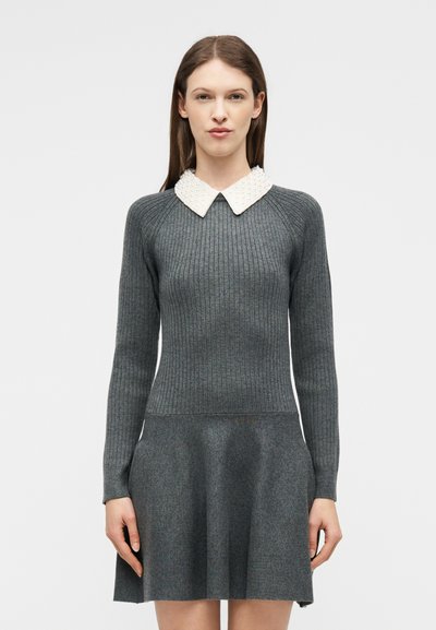 Femme portant une robe en tricot gris cintrée à rayures avec une jupe évasée et un col pointu texturé blanc, debout devant un fond uni.