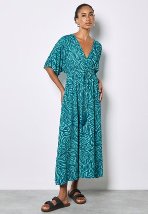 Frau steht mit Händen in den Taschen, trägt ein tealfarbenes maxikleid mit Muster und V-Ausschnitt sowie schwarze Sandalen vor einem einfarbigen hellen Hintergrund.