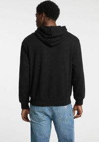 Felpa nera con cappuccio, dalla texture morbida, con polsini e vita a coste. Indossata con jeans azzurri, presenta un design pulito e semplice.