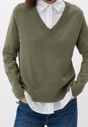 Pull en v olive avec des poignets et un ourlet côtelés, superposé sur une chemise blanche à col, offrant une coupe décontractée et une texture tricotée.