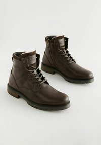 Botas de tobillo de cuero marrón con un acabado suave, diseño con cordones, cremallera lateral y suela de goma. Forro interior acolchado y detalle en el puño.