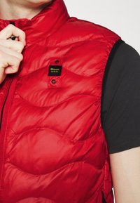 Gros plan sur une personne portant un gilet matelassé rouge par-dessus un t-shirt noir à manches courtes, la main ajustant la fermeture éclair du gilet.