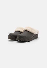 Bruna träskor med en mjuk krämfärgad fuskpälskant, rund tå och texturerad sula. Subtil design, lämplig för inomhus- eller avslappnad utomhusbruk.