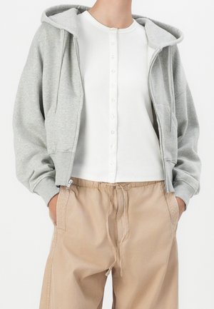 Osoba ubrana w jasnoszary bluzę z kapturem na zamek, na białą, prążkowaną bluzkę na guziki oraz beżowe spodnie ze sznurkiem, z rękami w kieszeniach.