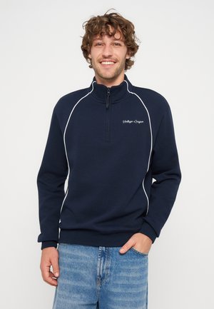 Jeune homme aux cheveux bouclés souriant, portant un sweat zippé bleu marine avec des passepoils blancs et un jean bleu clair.