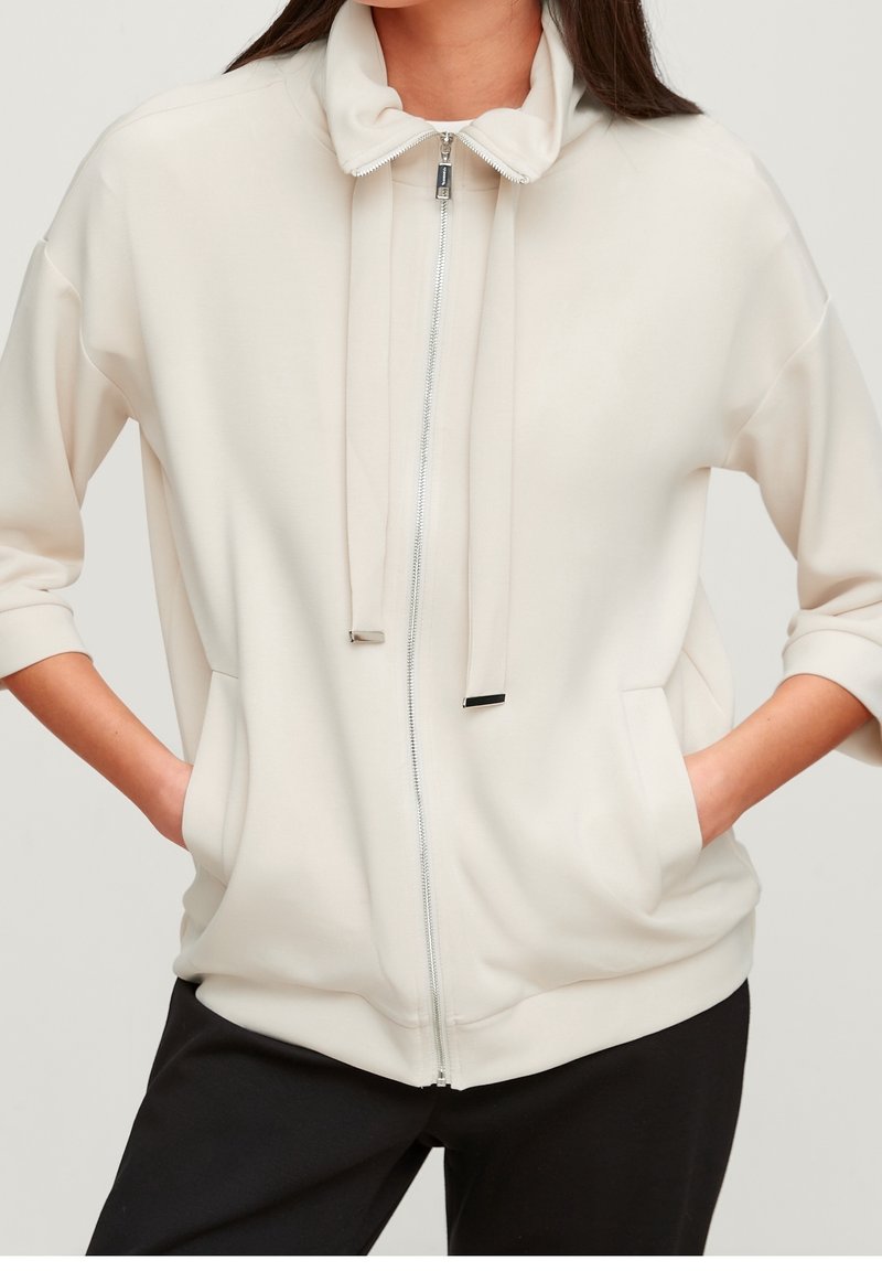 Femme portant une veste zippée écrue à col montant, avec cordons, manches trois-quarts et les mains dans les poches, associée à un pantalon noir.