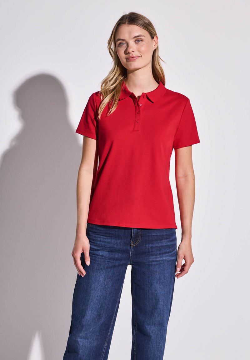 Polo rouge avec un col classique, des manches courtes et une patte de boutonnage à trois boutons, associé à un jean large bleu foncé. Tissu lisse et coupe élégante.