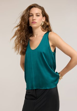 Femme aux longs cheveux ondulés porte un top en satin sans manches couleur sarcelle, un pantalon noir, des boucles d’oreilles dorées et un bracelet en or, debout, une main sur la hanche.
