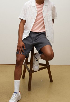 Homme assis sur un tabouret en bois portant une chemise blanche à manches courtes, un t-shirt rose, un short gris, des chaussettes blanches et des baskets blanches.