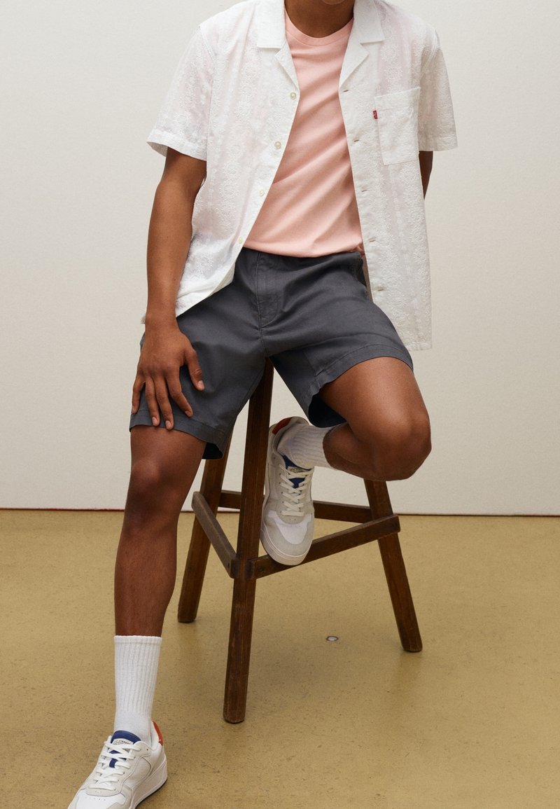Homme assis sur un tabouret en bois portant une chemise blanche à manches courtes, un t-shirt rose, un short gris, des chaussettes blanches et des baskets blanches.