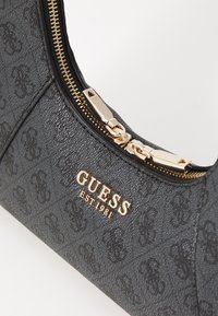 Sivá monogramová kabelka vyrobená z textúrovaného materiálu, s kultivovaným zlatým zipsom a kovovými doplnkami, pričom názov značky "GUESS EST. 1981" je výrazne zobrazený.
