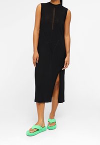 Robe noire sans manches en maille côtelée avec fente latérale, présentant un motif triangulaire à l'avant. Associée à des tongs vertes à semelle épaisse.