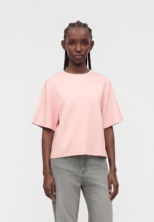Frau mit geflochtenem Haar trägt ein hellrosa kurzärmeliges Shirt und graue Jeans, steht vor einem schlichten hellgrauen Hintergrund.