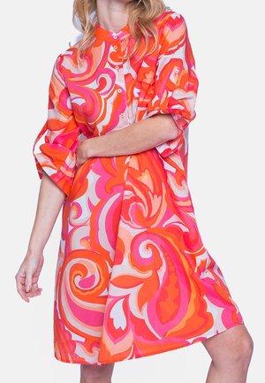 Kvinde iført en livlig knælang kjole med opkrogede ærmer, med dristige orange, pink og hvide swirl-mønstre.