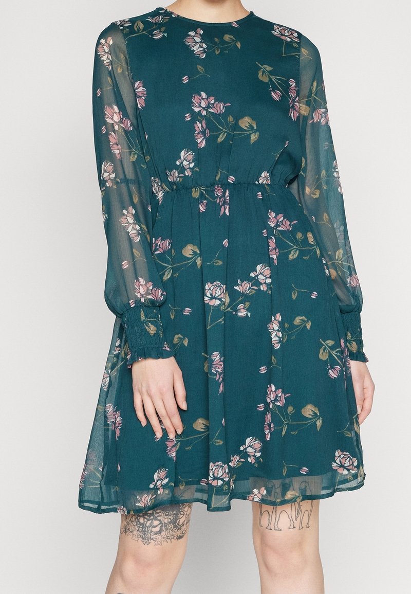 Robe bleu sarcelle à motif floral avec manches longues en voile transparent, taille froncée et jupe évasée. Présente des motifs botaniques roses et verts.