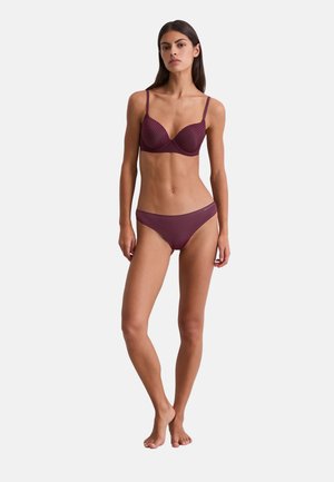 Vrouw staat op blote voeten en draagt een bijpassende donkerpaarse gestreepte beha en slip tegen een effen witte achtergrond.