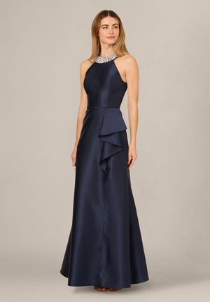Frau trägt ein ärmelloses marineblaues Satinkleid mit juwelenbesetztem Neckholder-Ausschnitt und gestuftem Volant-Detail an der Seite, steht vor einem schlichten Hintergrund.