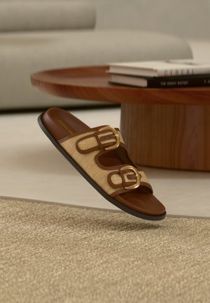 Sandale à talon marron avec deux boucles dorées sur des lanières tissées beige, flottant au-dessus d'un tapis beige texturé dans un salon moderne.