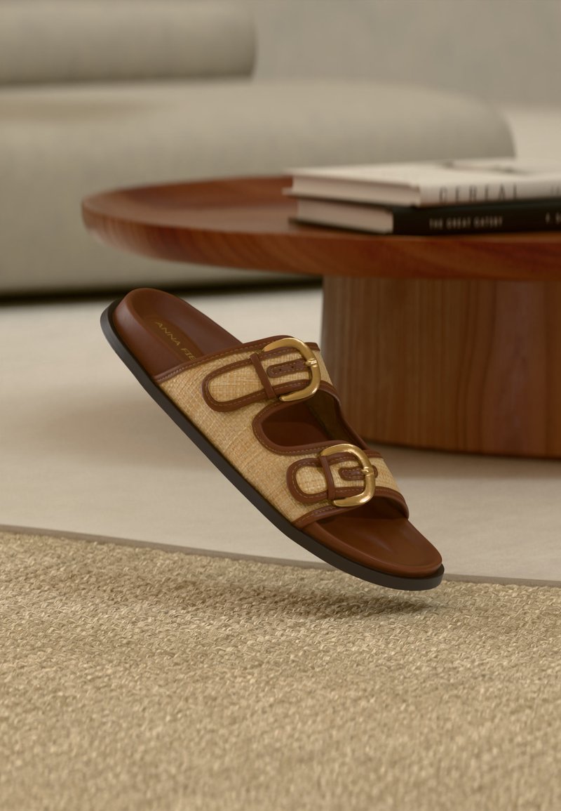 Sandale à talon marron avec deux boucles dorées sur des lanières tissées beige, flottant au-dessus d'un tapis beige texturé dans un salon moderne.