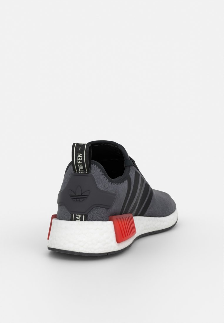 Grauer Sportschuh mit Obermaterial aus Stoff, drei schwarzen Streifen, rotem Heel-Akzent und weißer gepolsterter Sohle. Verfügt über eine Zuglasche und Markeninformationen.