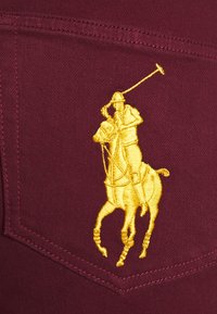 Tela burdeos con un acabado texturizado presenta un gran logo bordado de un jugador de pólo amarillo, detallado con un caballo y jinete en movimiento.