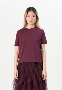 Young woman with long blonde hair wearing a plain dark purple short-sleeve shirt and layered ruffled dark purple skirt standing against white background.
