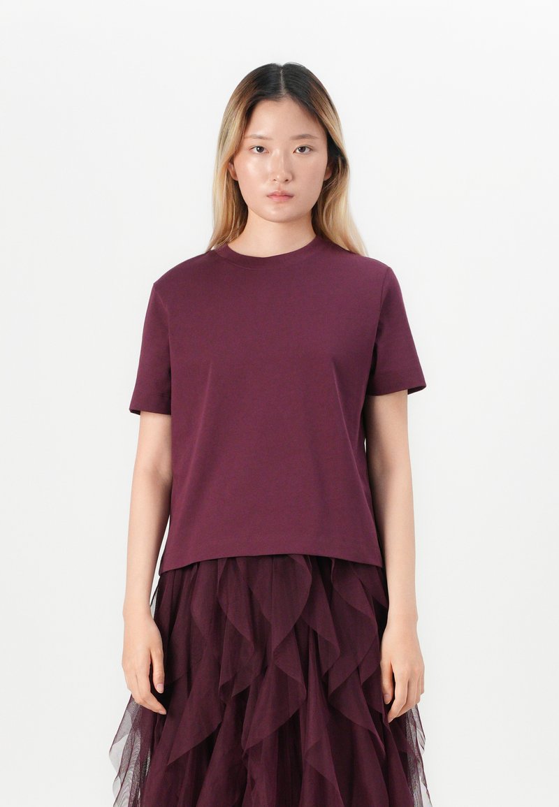 Young woman with long blonde hair wearing a plain dark purple short-sleeve shirt and layered ruffled dark purple skirt standing against white background.