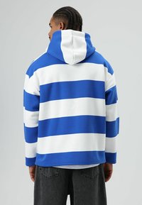 Blauw-wit gestreepte hoodie met een kangoeroezak en een capuchon met trekkoord, gemaakt van zacht materiaal. Draagt bijpassende donkergrijze broek.