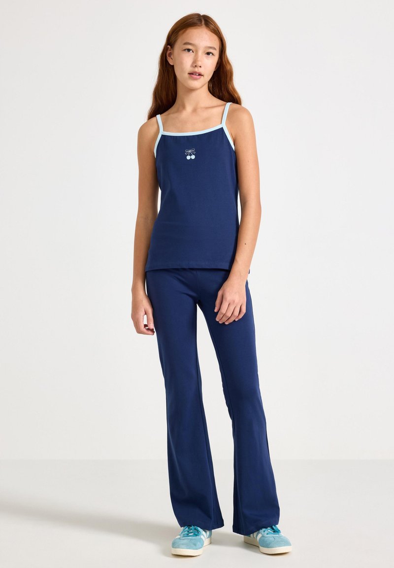 Marineblaues Tanktop mit hellblauen Trägern und Kirschmotiv, kombiniert mit marineblauen ausgestellten Hosen, getragen mit aquafarbenen Sneakern.