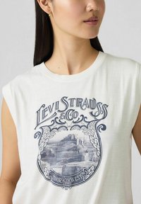 Biały bezrękawnik z niebieskim wzorem graficznym, zawierający napis "Levi Strauss & Co" oraz ilustrację górskiego krajobrazu.