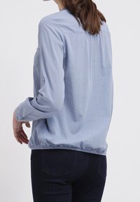 Chemise bleu clair à manches longues avec un tissu texturé, une coupe décontractée et un ourlet élastique. Comprend un empiècement dans le dos et une poche poitrine unique.