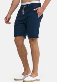 Granatowe szorty z wiązaniem w pasie, dwoma bocznymi kieszeniami i teksturowanym materiałem. Noszone z białymi espadrylami.