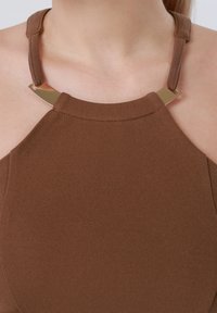 Καφέ τοπ με halter λαιμόκοψη και χρυσές μεταλλικές λεπτομέρειες στα λουριά, φορεμένο από άτομο με εμφανή λαιμό και ώμους.