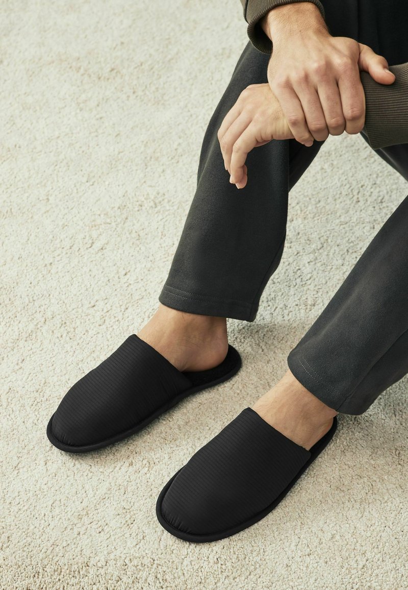 Chaussons noirs à enfiler avec une texture côtelée et un design rembourré. Doublure intérieure douce et semelle plate et flexible. Portés avec un pantalon sombre.