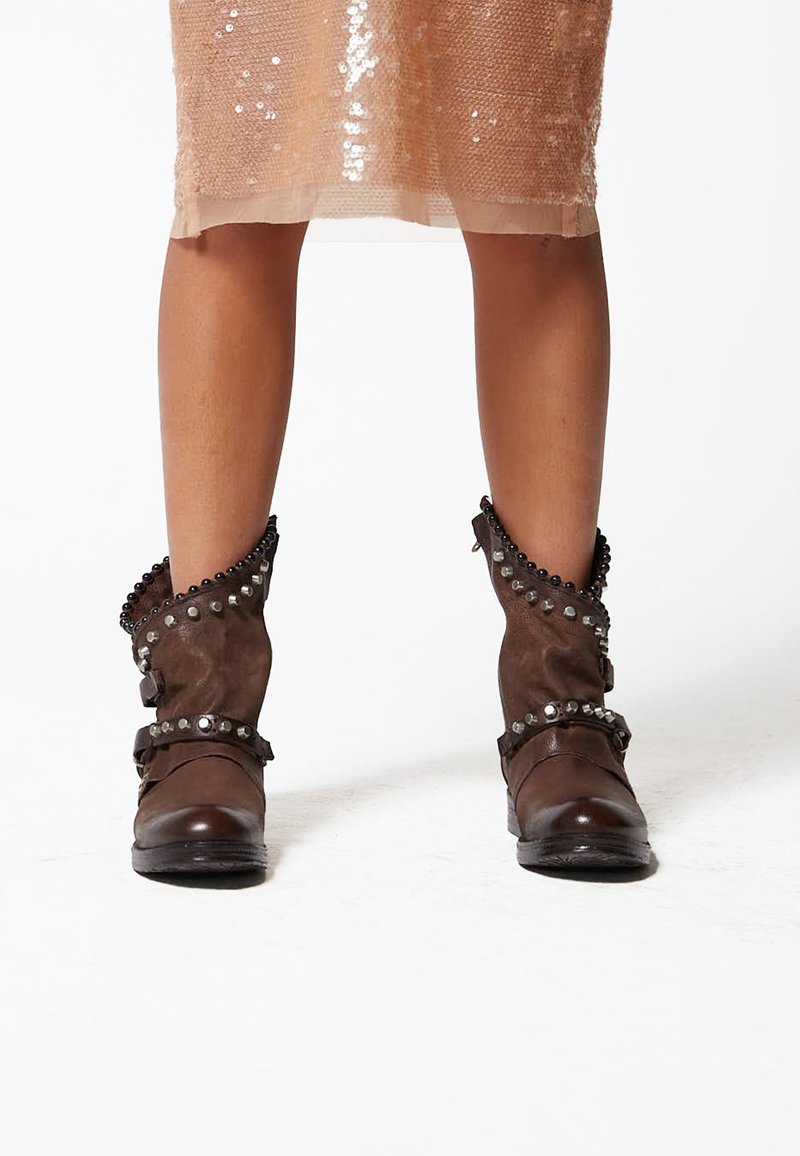 Brown leather ankle boots with studded accents and a textured fabric cuff. Features straps and a rounded toe design.